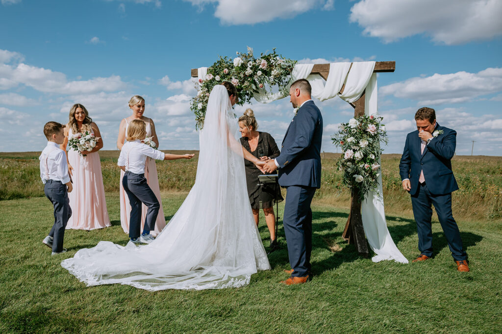 A wedding ceremony taking place outdoors with guests standing around the bride and groom under a floral arch.