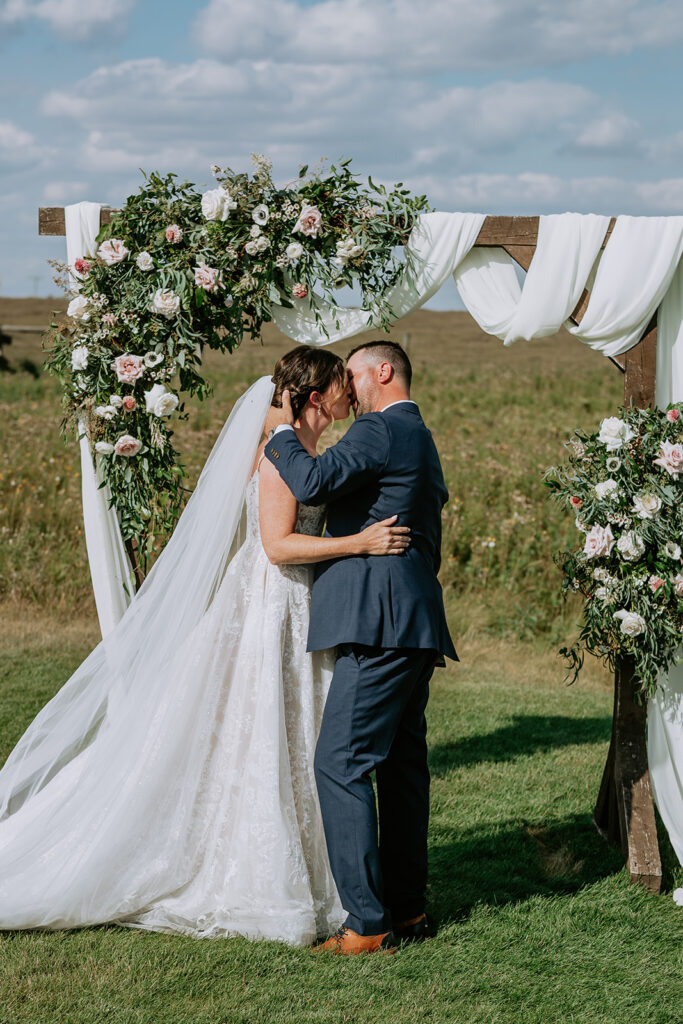 A bride and groom share a kiss under a floral archway at an outdoor wedding ceremony.