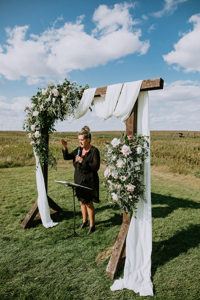 A woman speaking at a podium beside a floral-decorated arch at an outdoor event.