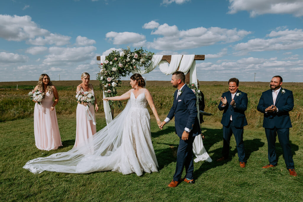 A bride and groom hold hands at their outdoor wedding ceremony, with bridesmaids in pink dresses and groomsmen clapping.