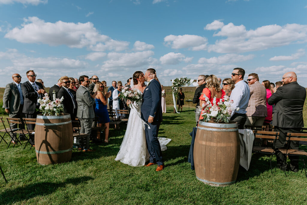 A bride and groom share a kiss as they walk down the aisle after their outdoor wedding ceremony, surrounded by guests and flanked by floral arrangements on wooden barrels.