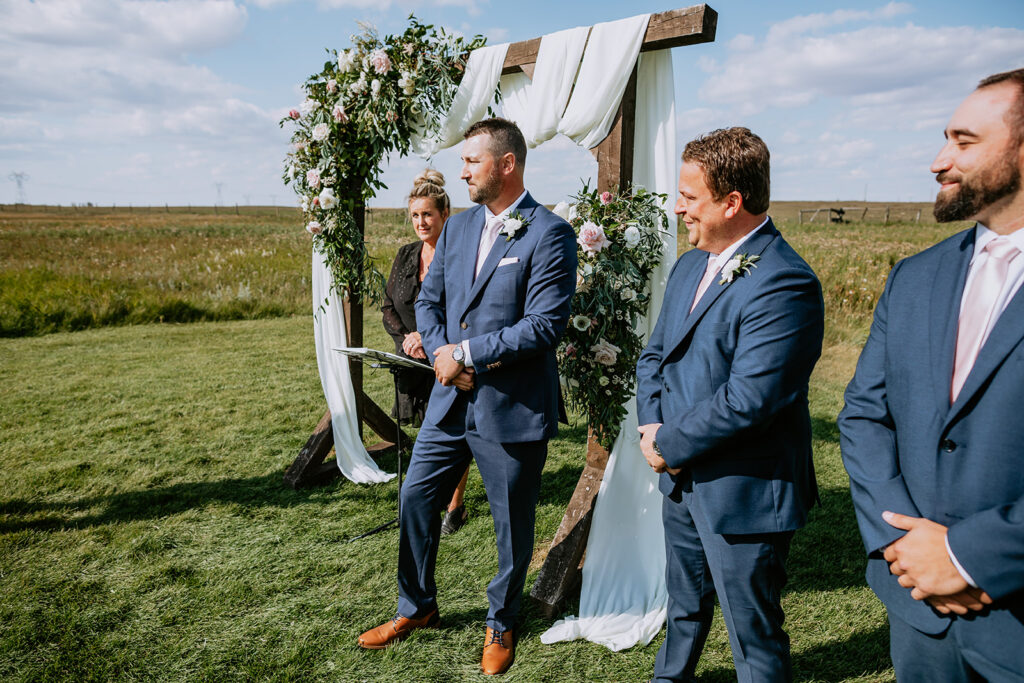 Groom standing with groomsmen at an outdoor wedding ceremony by a floral arch.