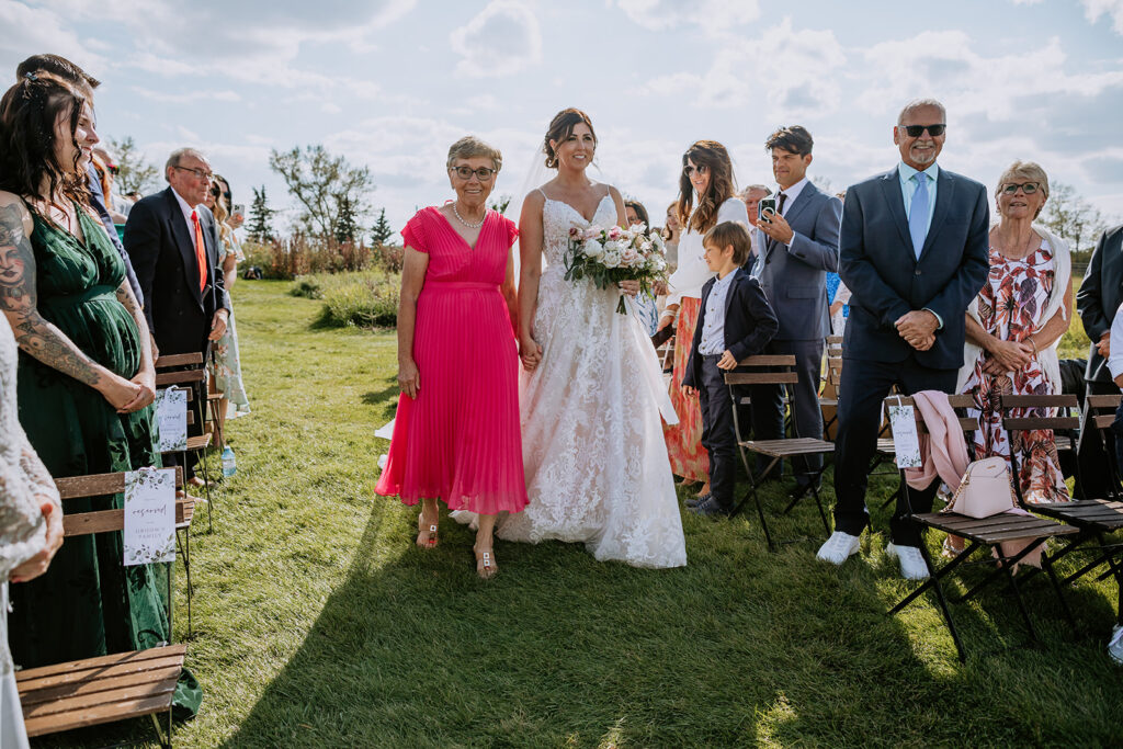 A bride walks down the aisle with an older woman by her side, while guests watch at an outdoor wedding ceremony.