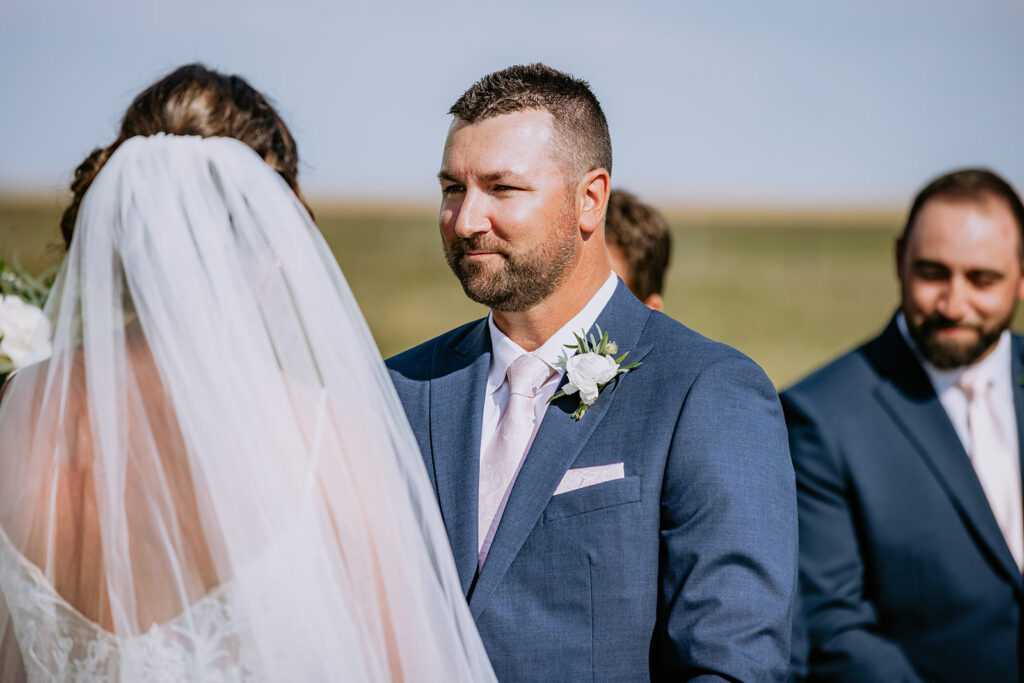A groom in a blue suit looking at his bride with emotion during their wedding ceremony, with a groomsman in the background.