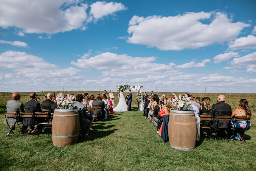 Outdoor wedding ceremony with guests seated on chairs facing a couple at the altar, under a clear blue sky.
