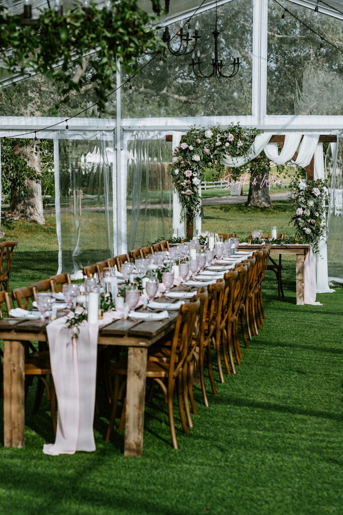 Elegant outdoor wedding reception setup with long wooden tables, floral centerpieces, and clear tent on a lawn.