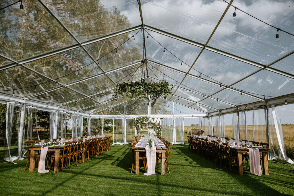 Elegant outdoor event setup inside a clear tent with long dining tables and suspended greenery decoration.