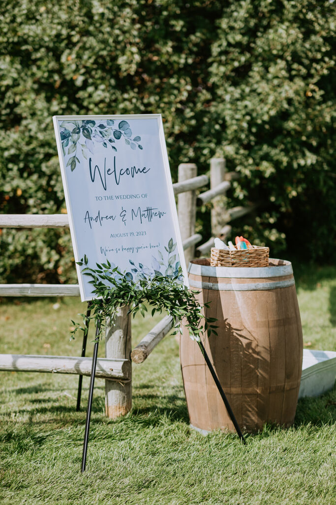 A welcome sign for a wedding, featuring the names of the bride and groom, placed on an easel beside a wooden barrel under sunny outdoor conditions.