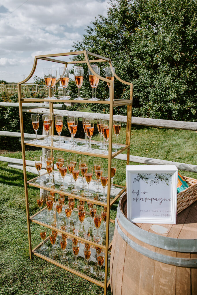An outdoor champagne glass display on a gold shelving unit with a "help yourself to champagne" sign and a wooden barrel nearby.