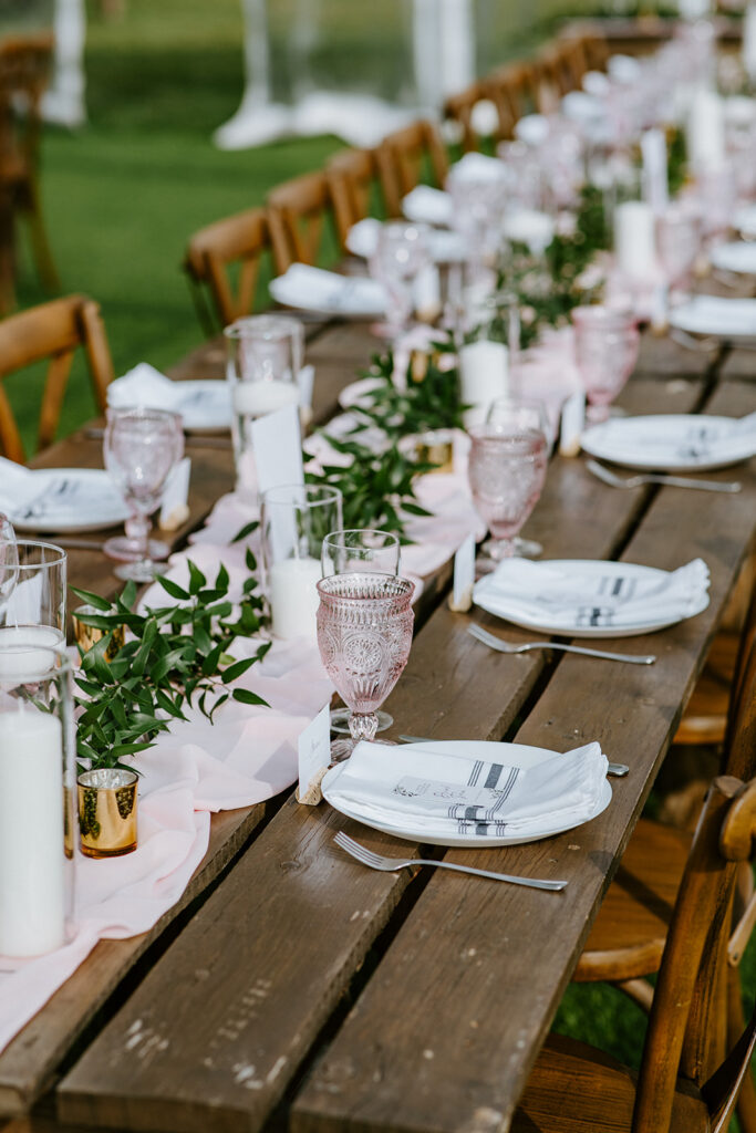 Elegant outdoor dining setup with a long wooden table, place settings, and decorative greenery.