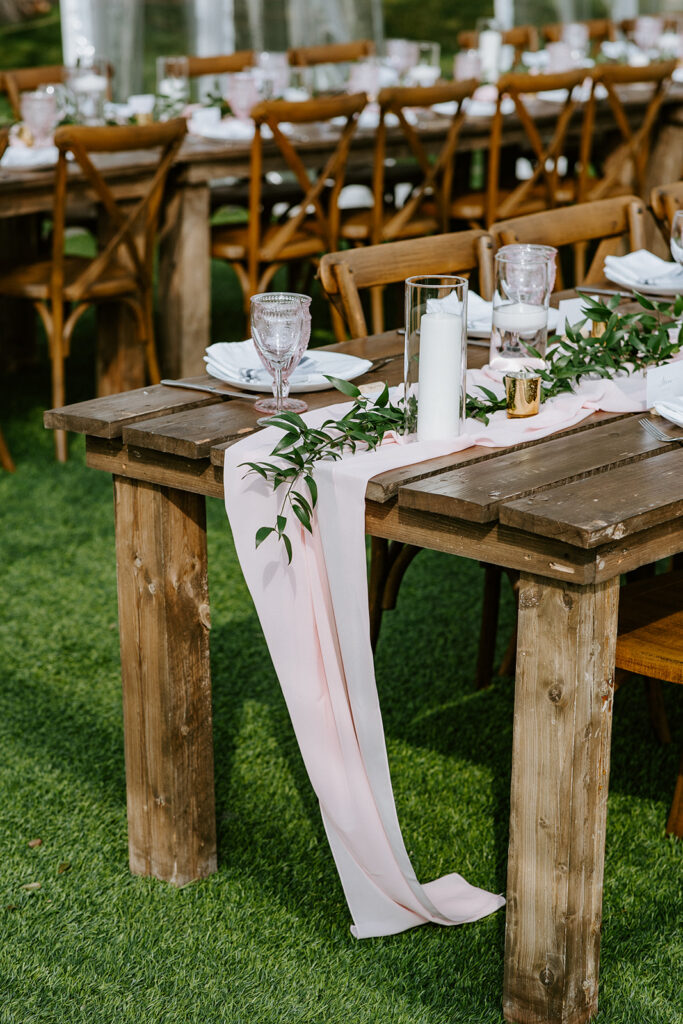 An elegantly set outdoor dining table with rustic chairs, greenery decorations, and a pink table runner.