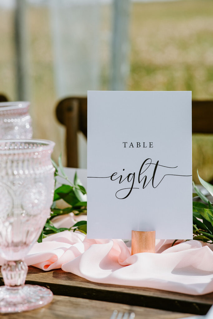 Elegant table number eight sign with cursive writing, set on a wedding reception table with decorative glassware and a pink ribbon.