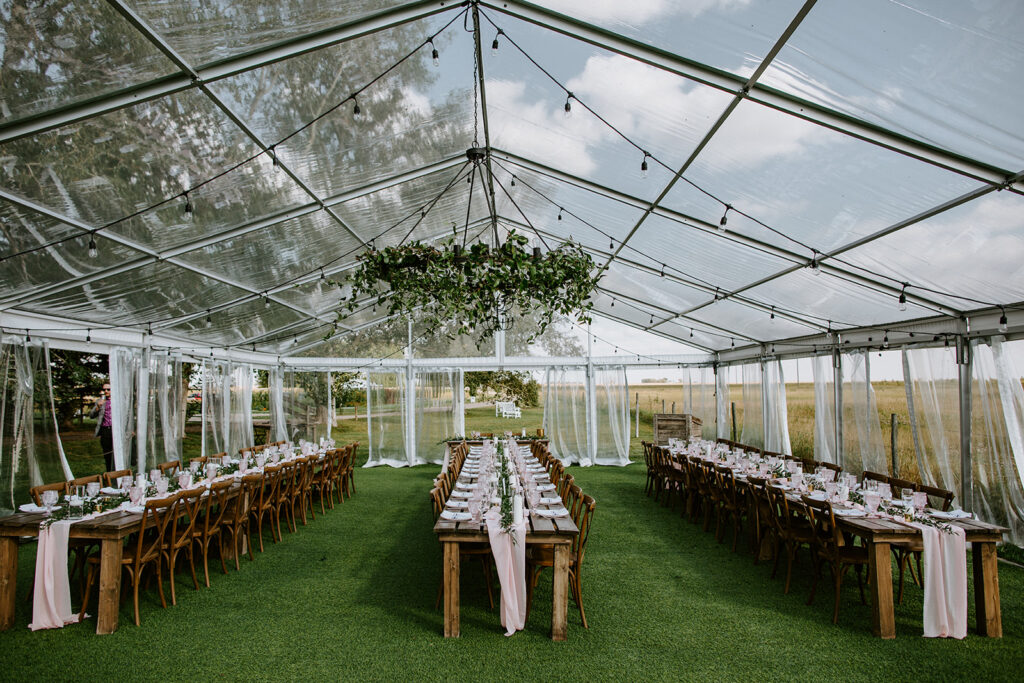 Elegant outdoor wedding reception setup under a clear tent with long wooden tables and a floral chandelier centerpiece.