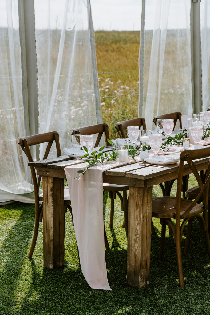 Elegant outdoor dining setup with wooden chairs and a table adorned with greenery and a pink runner, against a backdrop of sheer curtains and a natural field.