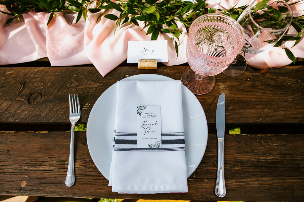 Elegant table setting for a wedding, complete with a personalized menu and a pink glass on a wooden table.