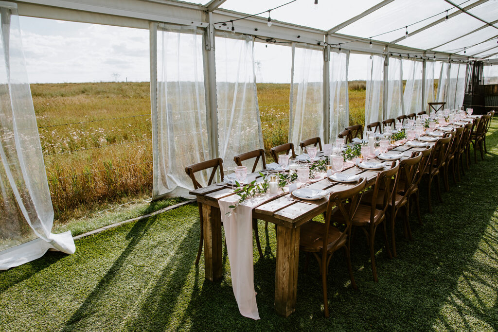 Outdoor banquet setup under a tent with wooden tables and elegant place settings, overlooking a field.