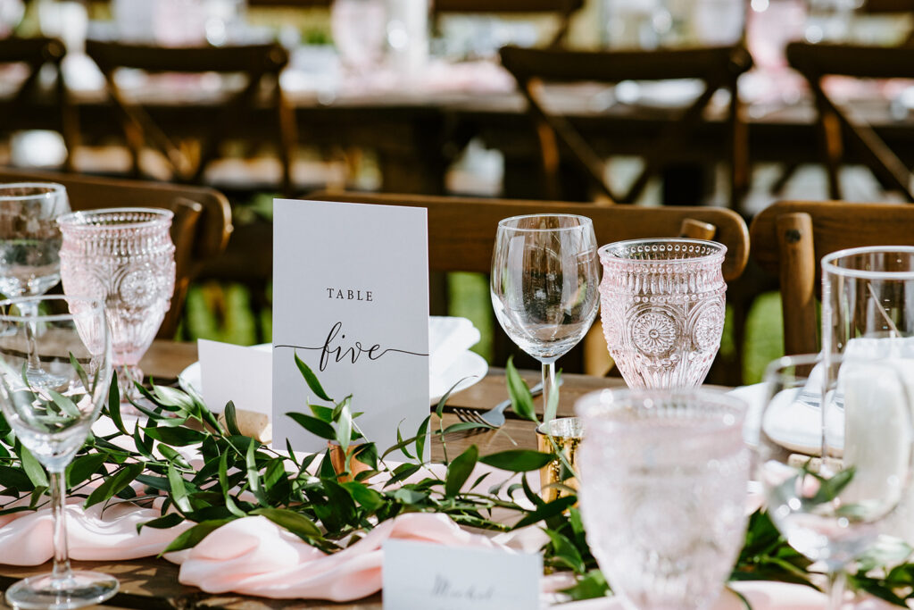 Elegant table setting for a formal event with a 'table five' sign, glassware, and decorative greenery.