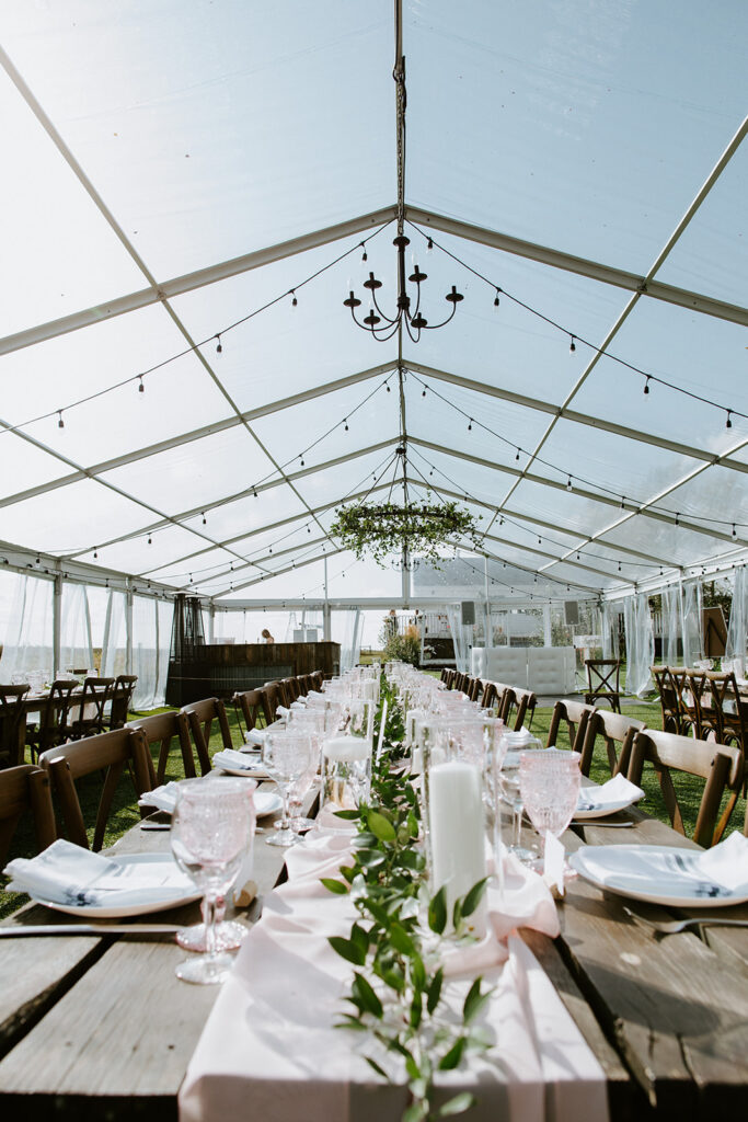 Elegant outdoor wedding reception setup with a clear tent, chandeliers, and decorated tables.