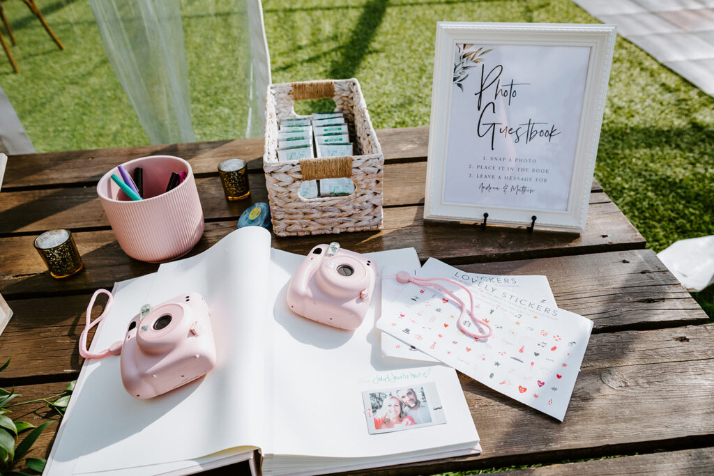 A photo guestbook station at an outdoor event with instant cameras, a sign, markers, and stickers for guests to use.