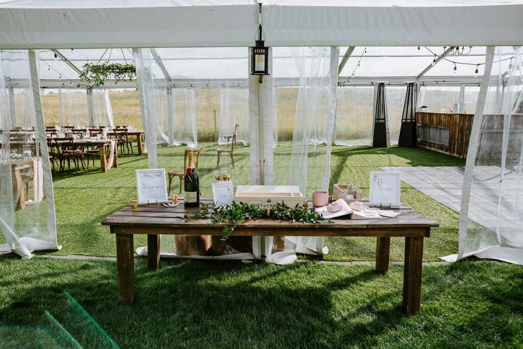 An outdoor wedding setup with a wooden table featuring a guest book and champagne under a tent with transparent curtains.