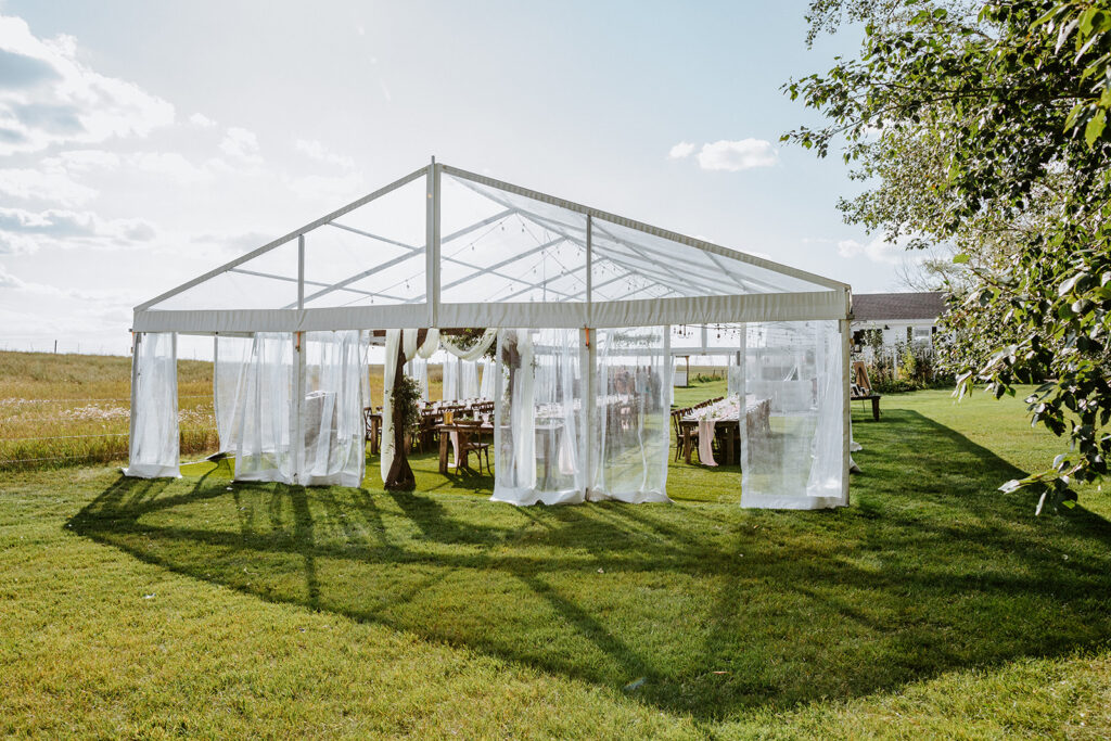 A transparent marquee set up for an event on a grassy field with open curtains and tables inside under a clear sky.