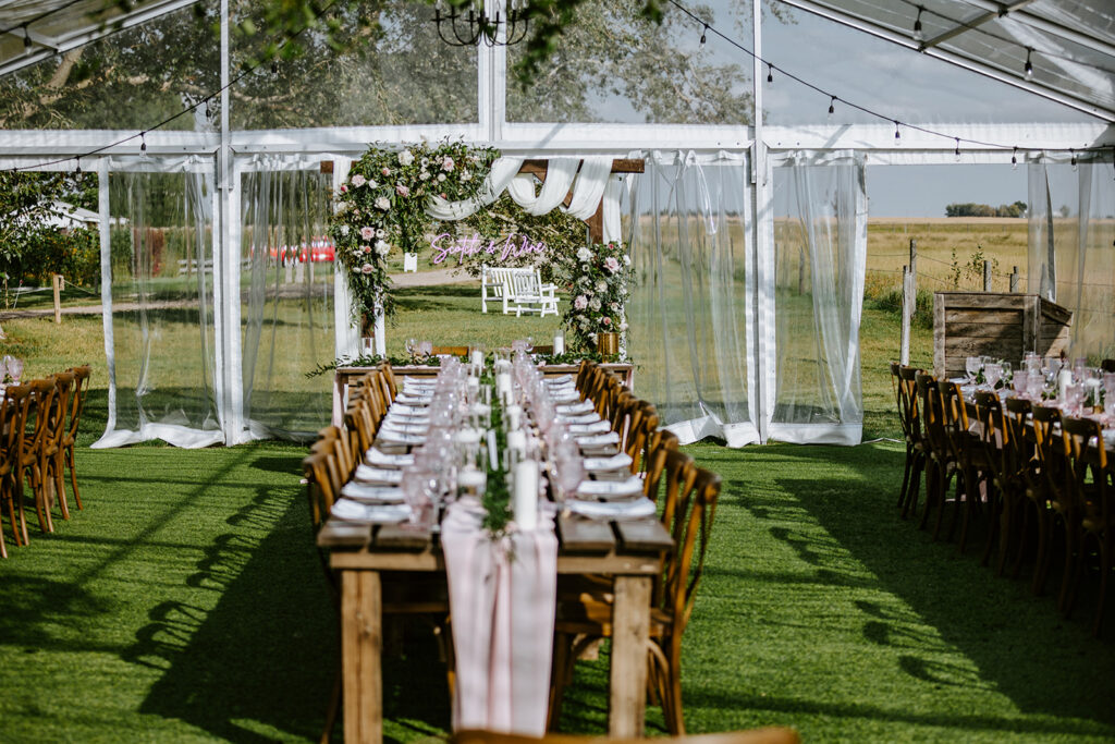 Outdoor wedding setting with a long dining table under a clear tent, adorned with floral arrangements and facing an altar.