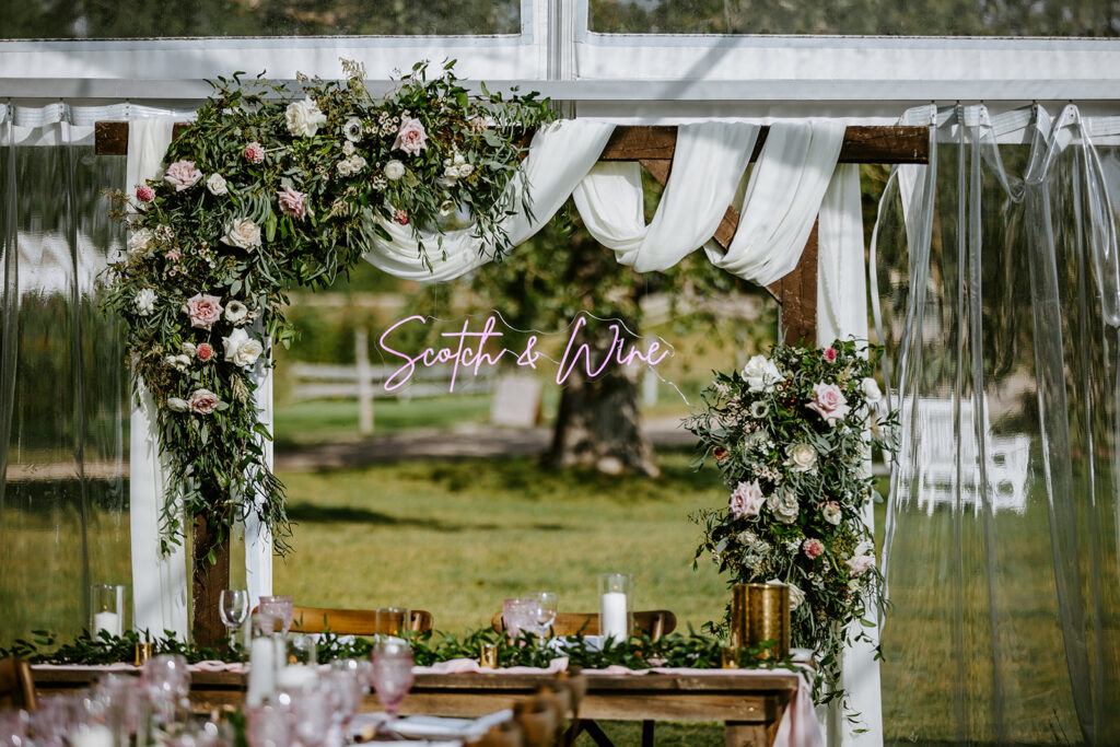 Elegant outdoor wedding setup with floral arch and "scott & kira" sign.