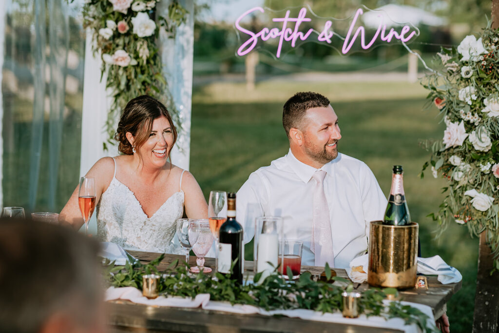 A smiling couple seated at a wedding table with decorative flowers and a sign reading "scotch & wine.