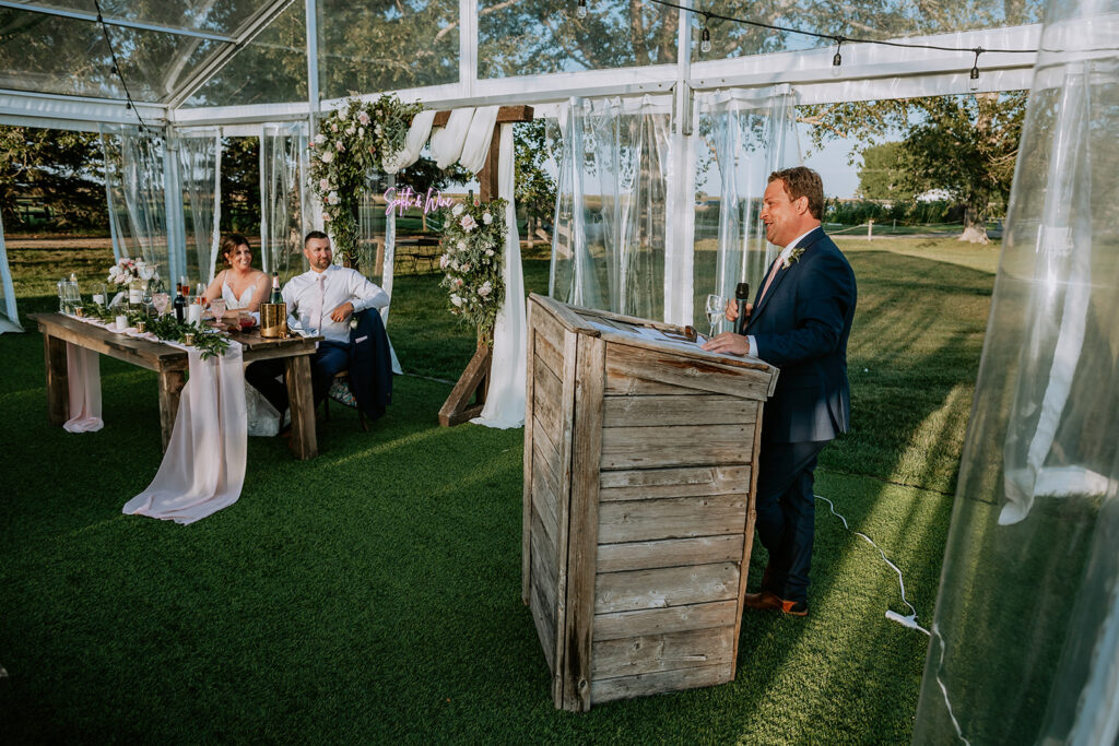 A man delivers a speech at an outdoor wedding reception while the bride and groom listen from their table.