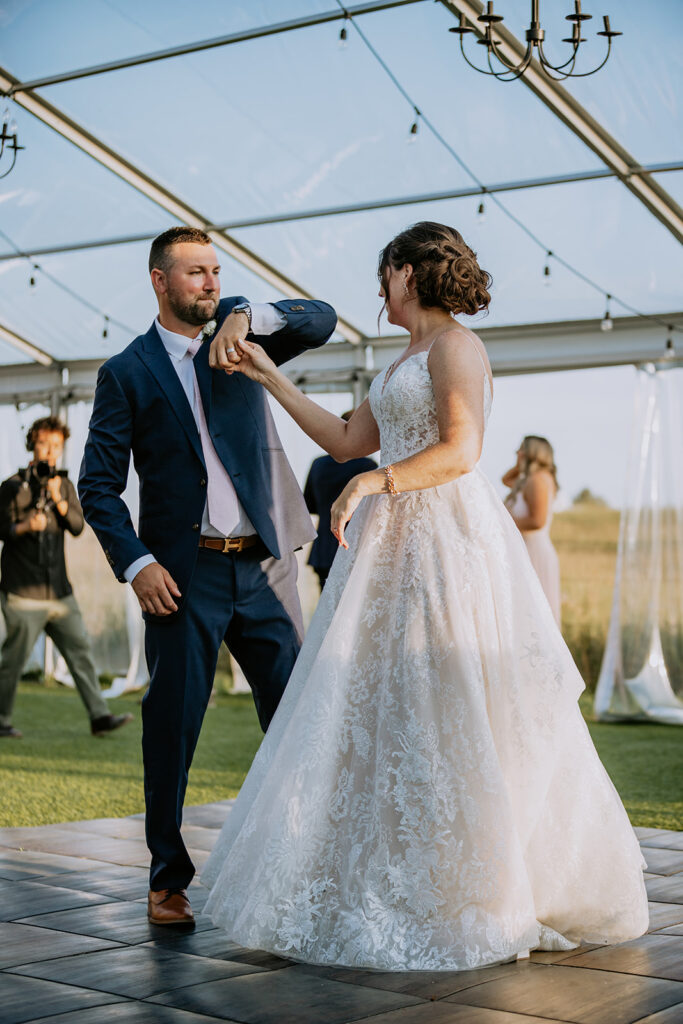 A couple in formal wedding attire dancing outdoors under a tent with a chandelier, with a photographer in the background.