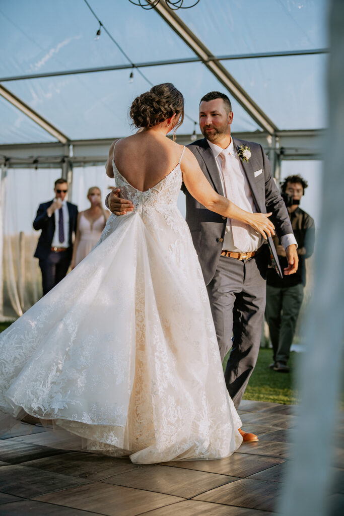 A bride and groom share a dance under a tent at their wedding, surrounded by guests.