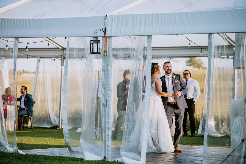 Bride and groom smiling during an outdoor wedding ceremony under a tent with guests looking on.
