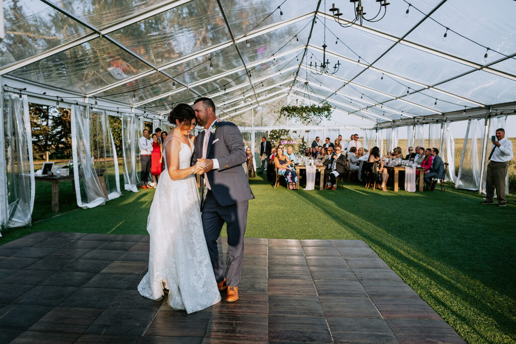 Bride and groom sharing a first dance under a transparent marquee with guests looking on.