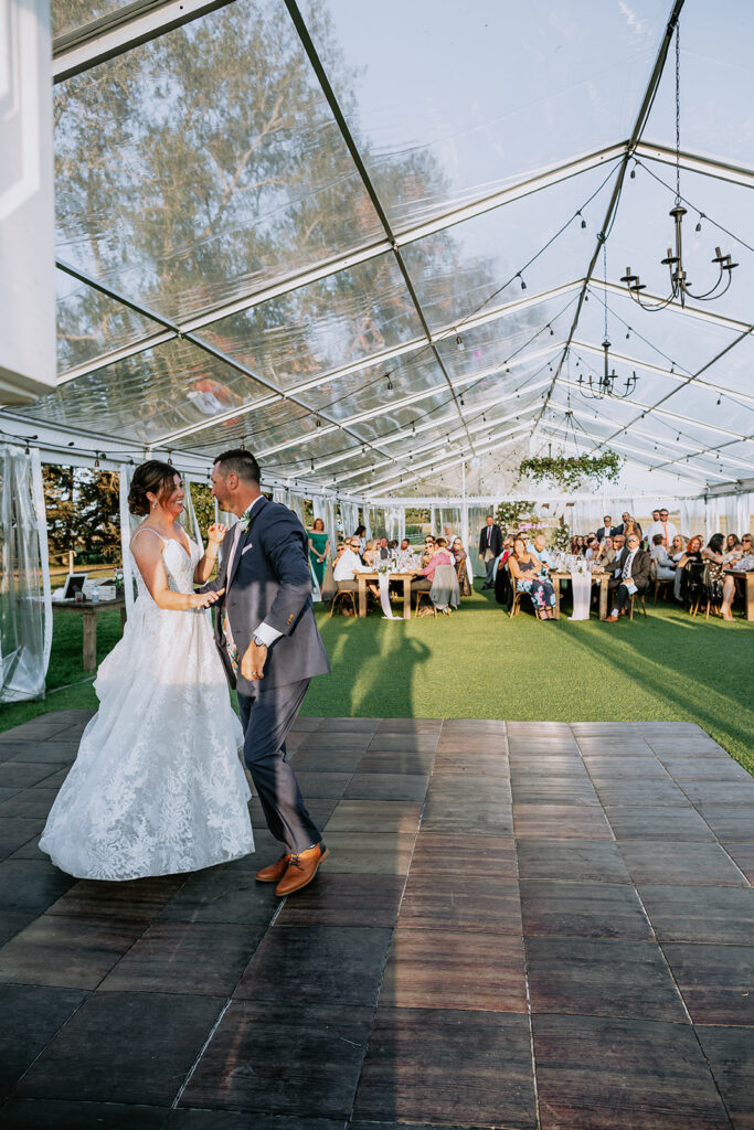 Bride and groom walking hand in hand on a dance floor under a tent at an outdoor wedding reception.