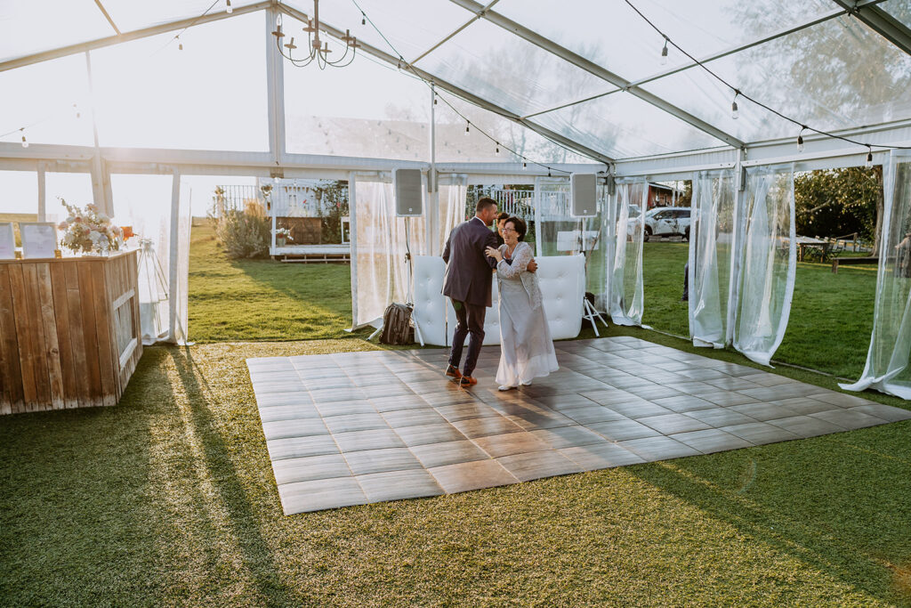A newlywed couple sharing a dance inside a tent with open curtains and a wooden floor at a daytime outdoor wedding reception.