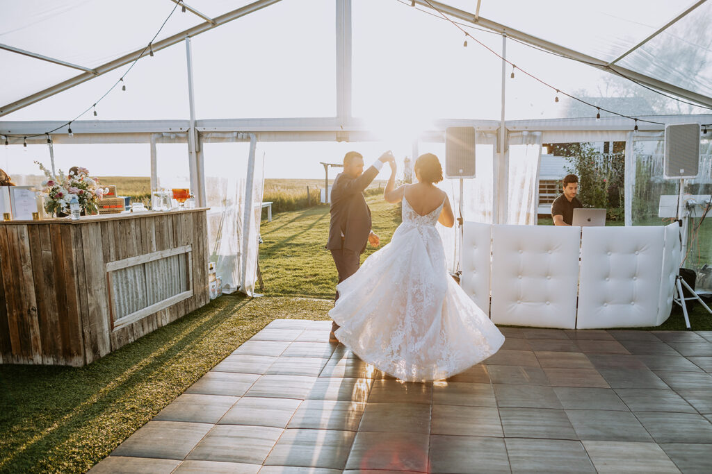 Bride and groom share a dance under a tent at sunset with a dj in the background.