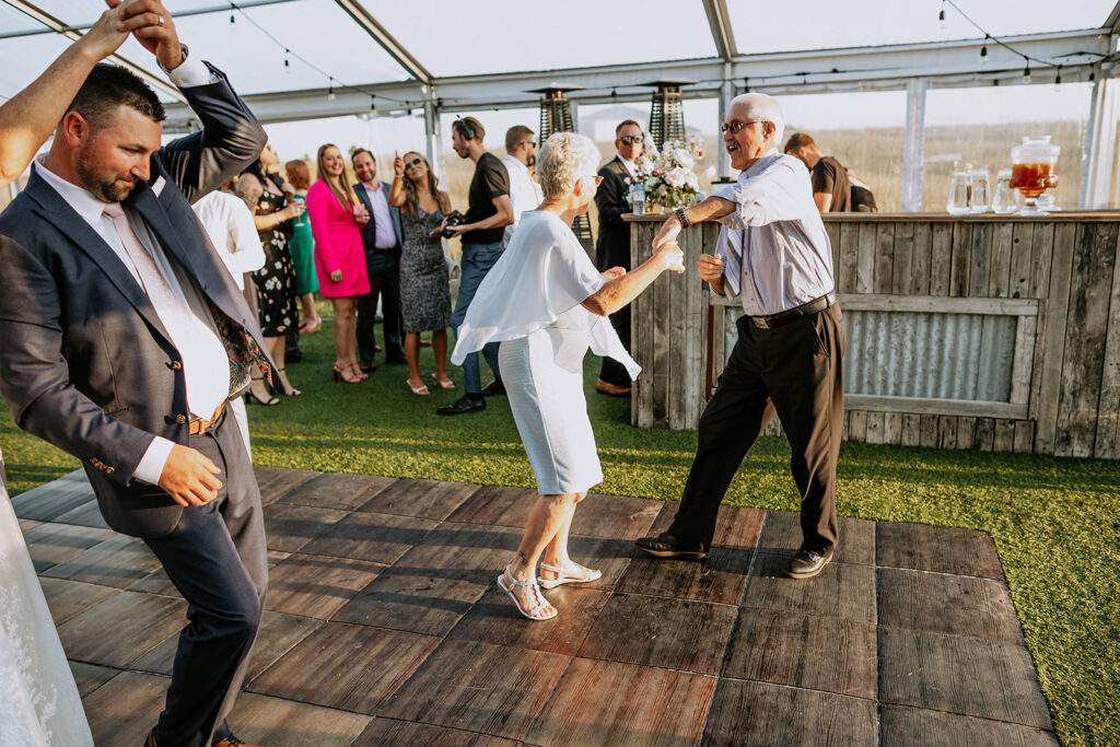 An elderly couple joyfully dancing together at an outdoor event while surrounded by smiling guests.