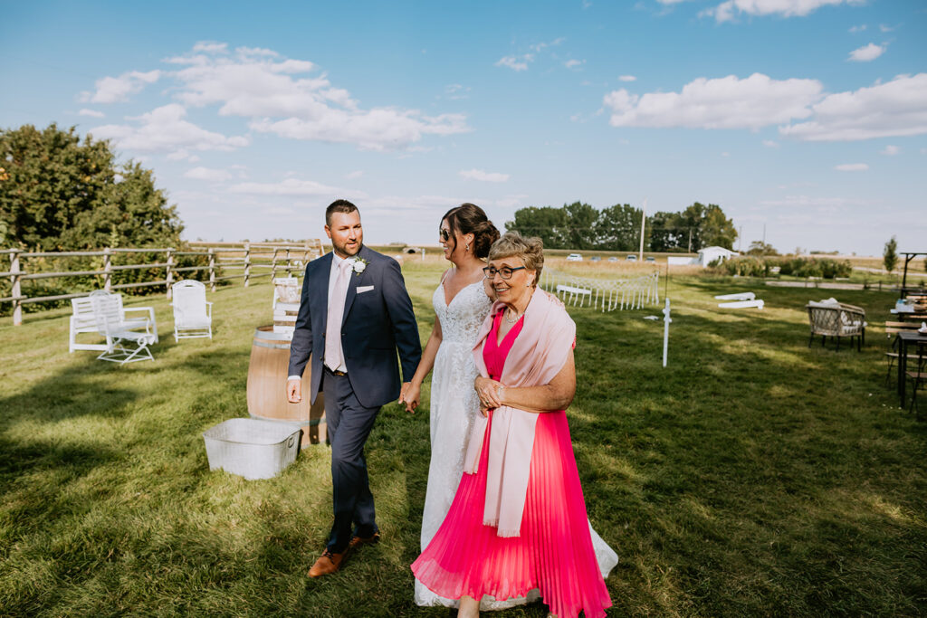 A bride accompanied by two individuals walks across a grassy area with chairs and a clear sky above.