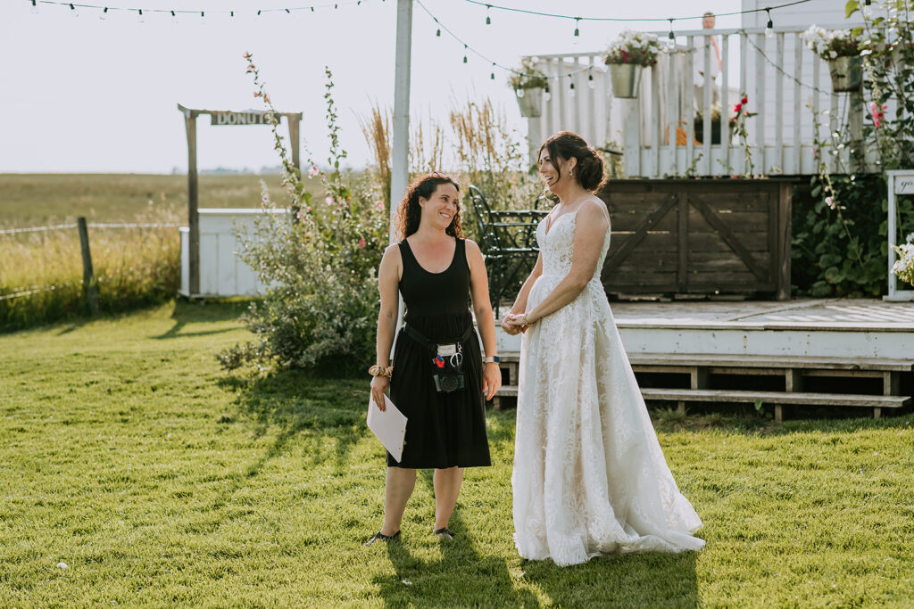 Two women smiling at each other, one in a wedding dress and the other in a black dress, standing on grass with festive decorations in the background.