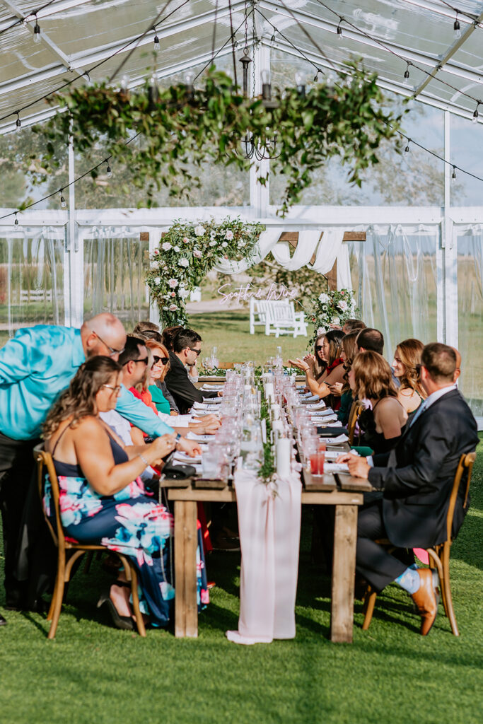 Guests seated at a long dining table under a clear tent at an outdoor event.