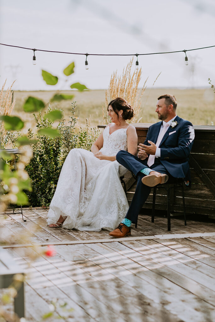 A bride and groom sitting side by side on a bench outdoors, looking relaxed and happy on their wedding day.