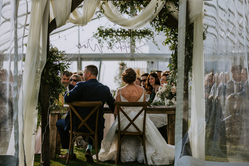 Bride and groom seated at their wedding table facing guests during a ceremony or reception.