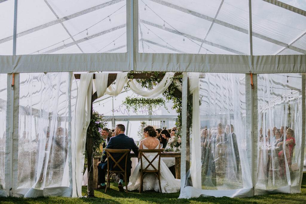 A wedding ceremony taking place inside a tent with open drapes, guests seated, and the couple sitting at the altar.