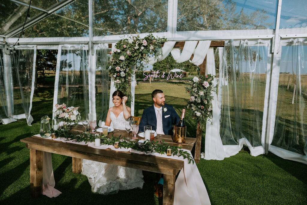 A bride and groom sit at a decorated wedding table under a floral arch, with the outdoors visible through a clear tent.