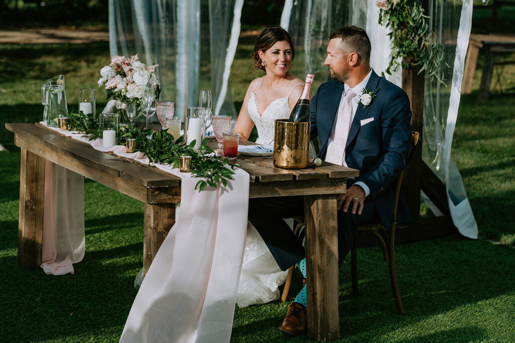Bride and groom sitting at a rustic wooden table during a wedding reception outdoors.