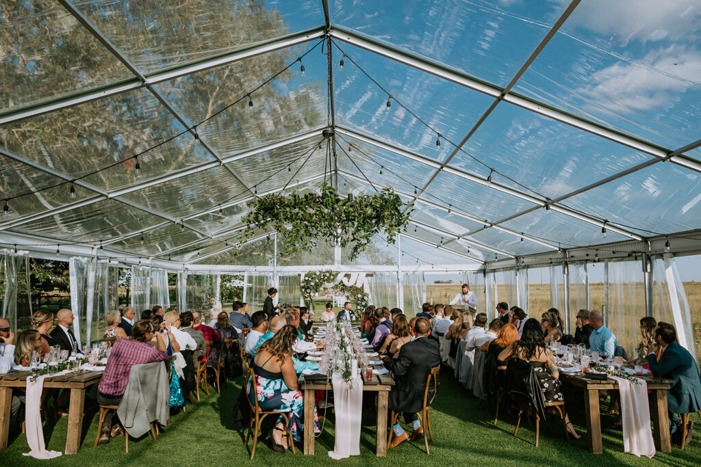 Guests seated at long tables during an event inside a clear tent with open countryside surroundings.