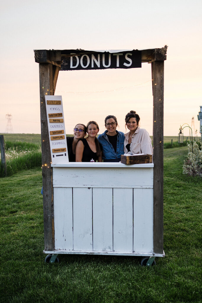 Four individuals posing behind a rustic outdoor donut stand at twilight.