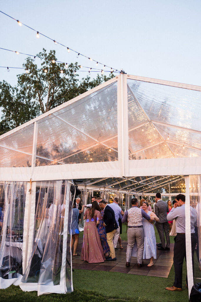 Guests dancing at an outdoor evening event under a clear tent adorned with string lights.