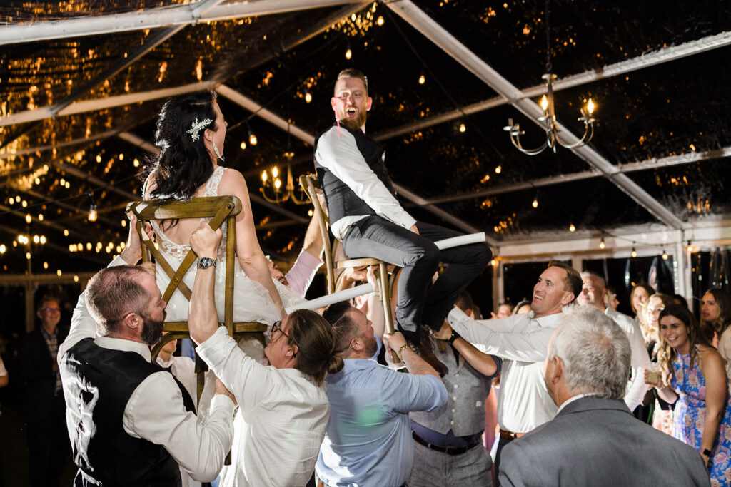 A bride and groom joyfully being lifted on chairs by guests in a traditional chair dance at a wedding reception.