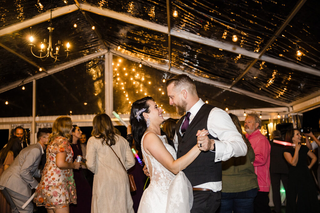 A bride and groom share a dance under a tent adorned with string lights, surrounded by wedding guests.